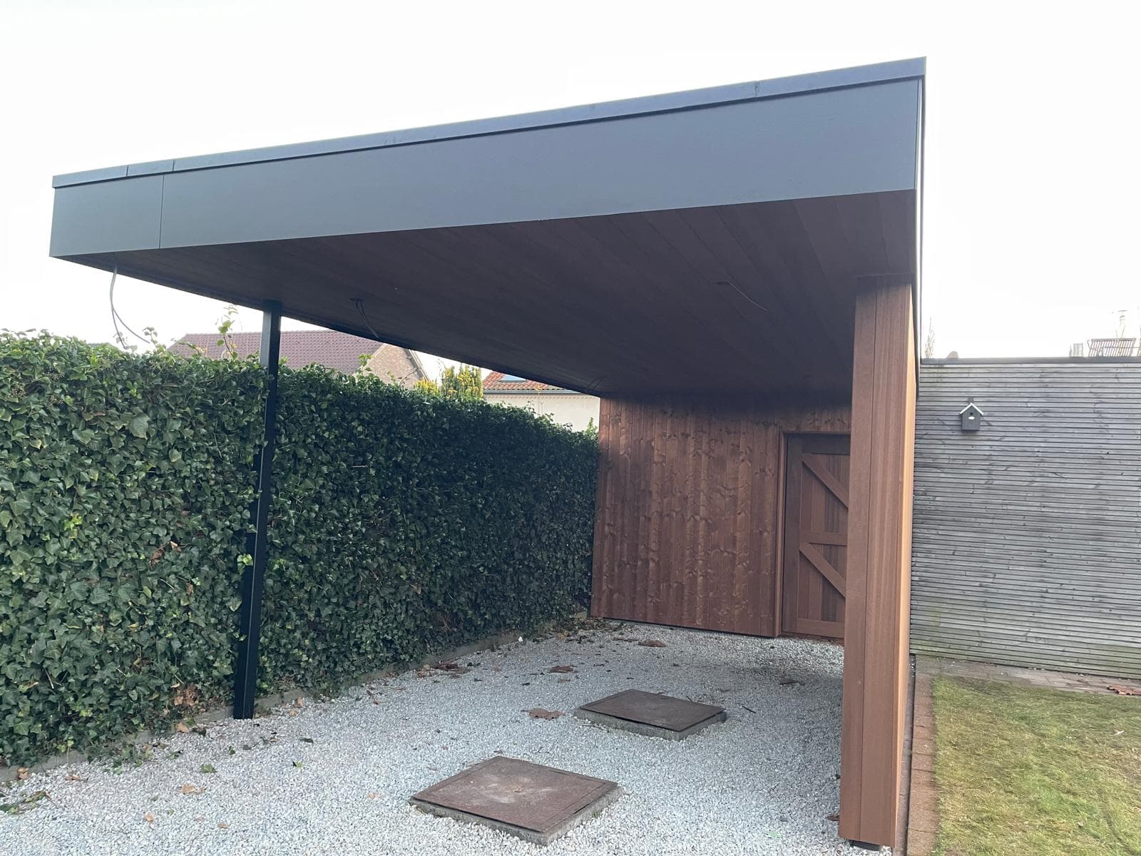 Zicht onder de carport met zwarte stalen kolom, houten plafond en grindvloer