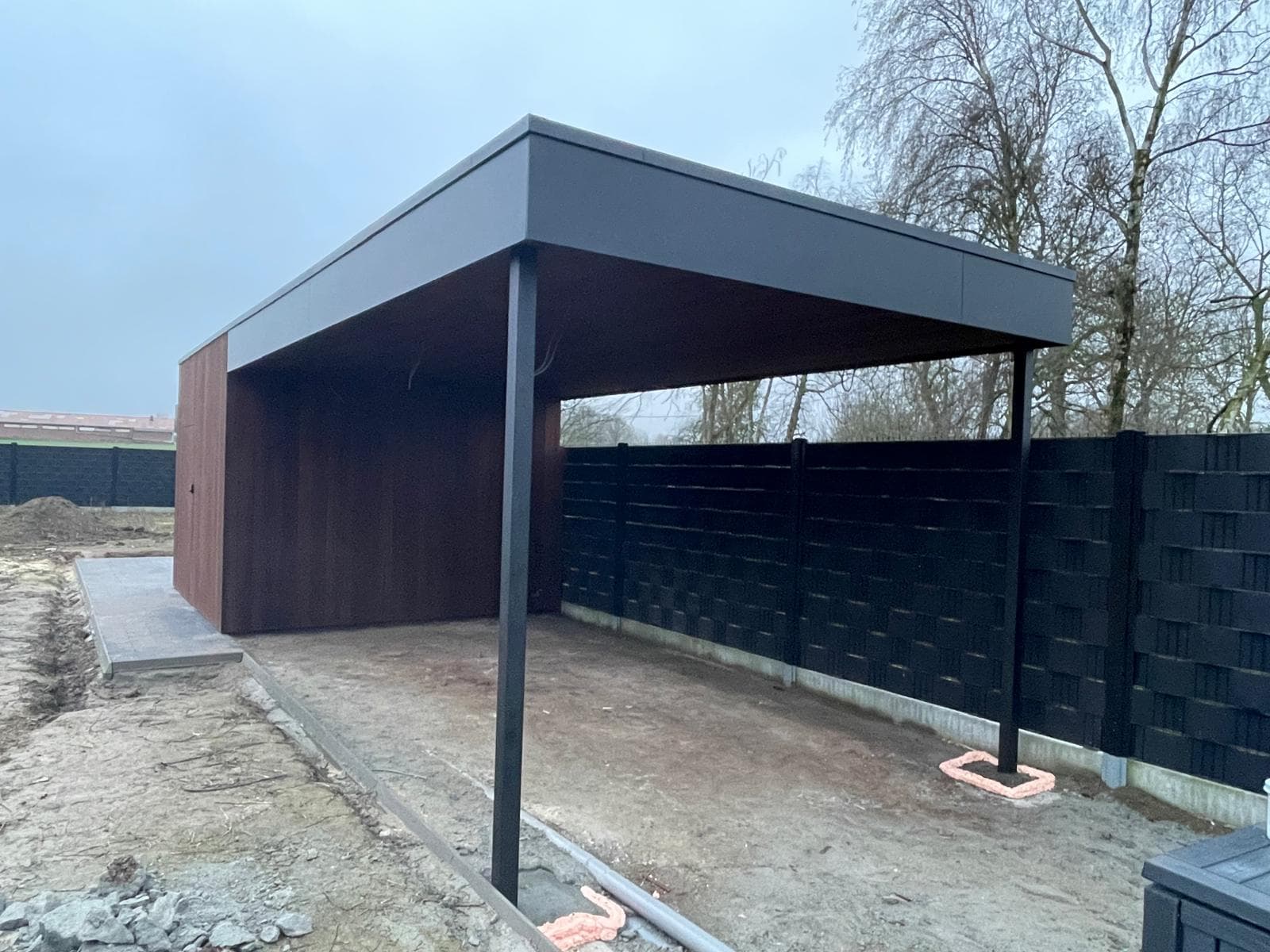 Carport met zwart metalen dak op slanke stalen palen langs een zwarte schutting
