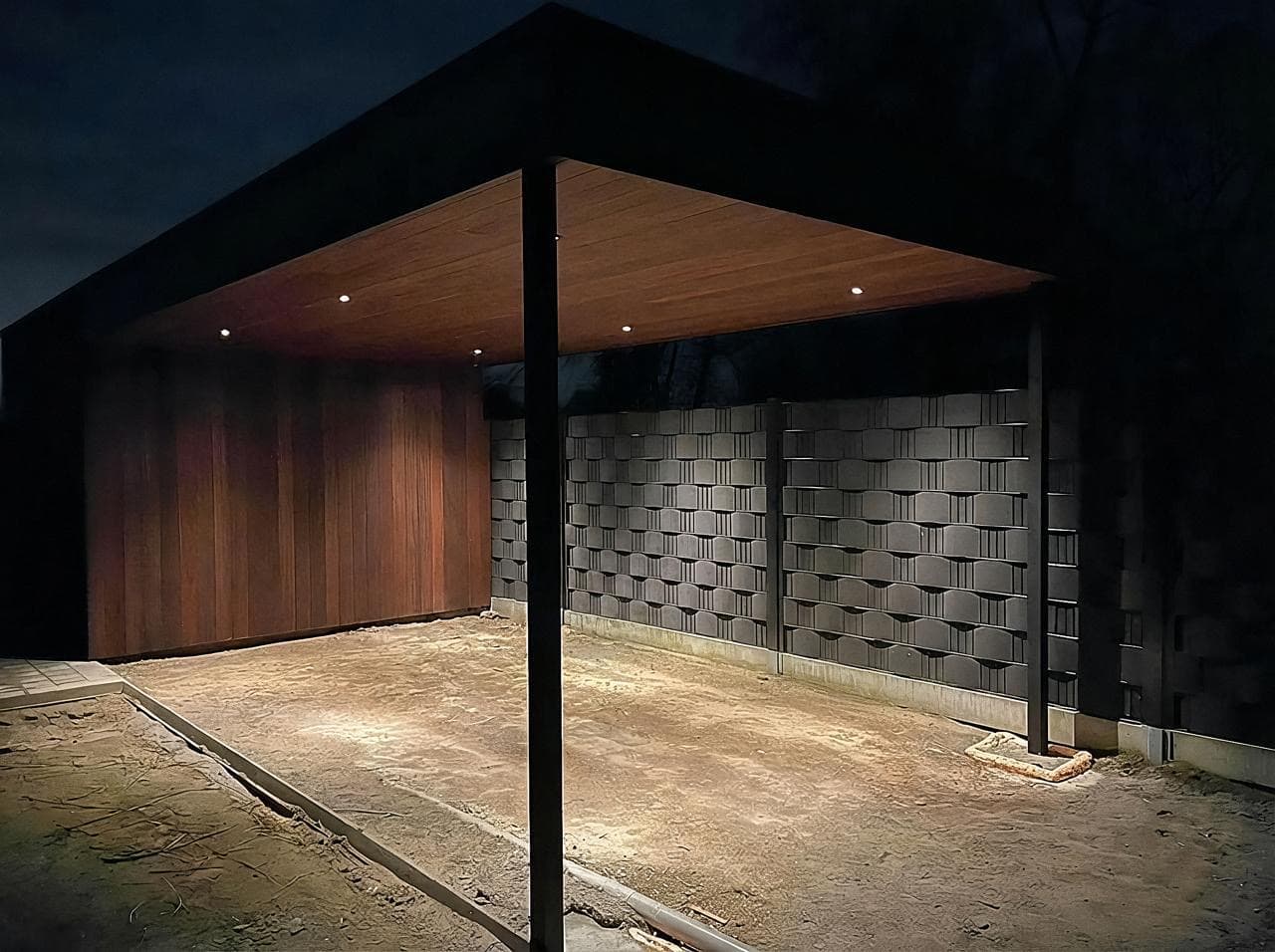 Carport met tuinberging in Ayous thermohout, sfeervol verlicht bij avond met zwarte trespa en metalen palen, WoPa Construction