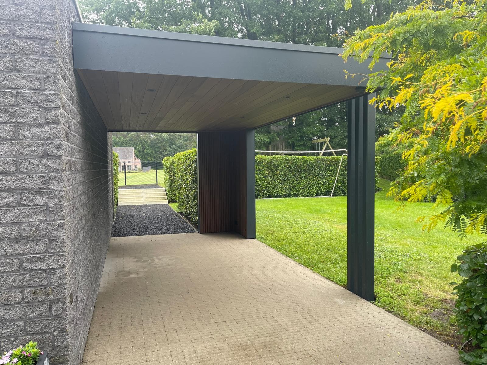 Doorkijk onder de carport langs de gevelsteenmuur met Ayous thermo houten plafond en zwarte stalen kolom