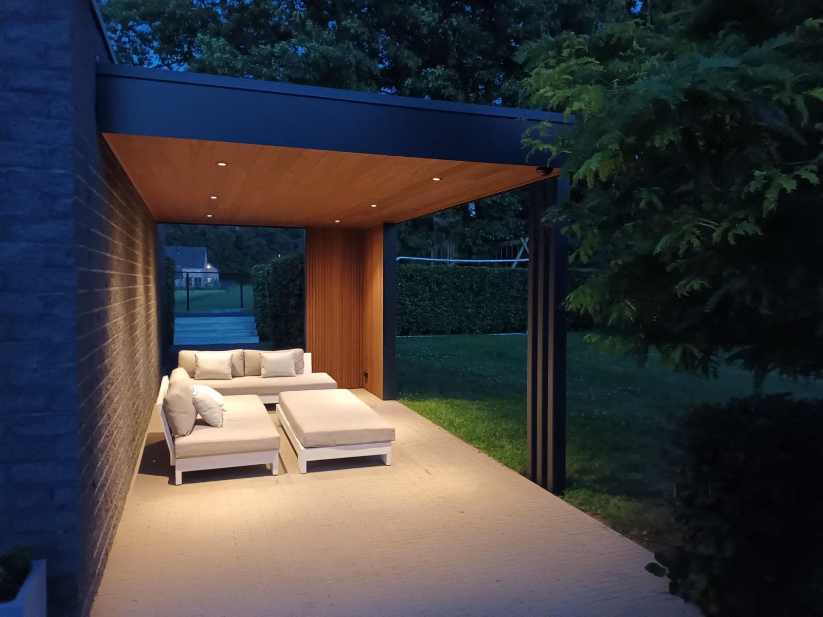 Avondsfeer met verlichte loungebanken onder het warm verlichte houten plafond en doorkijk naar de tuin