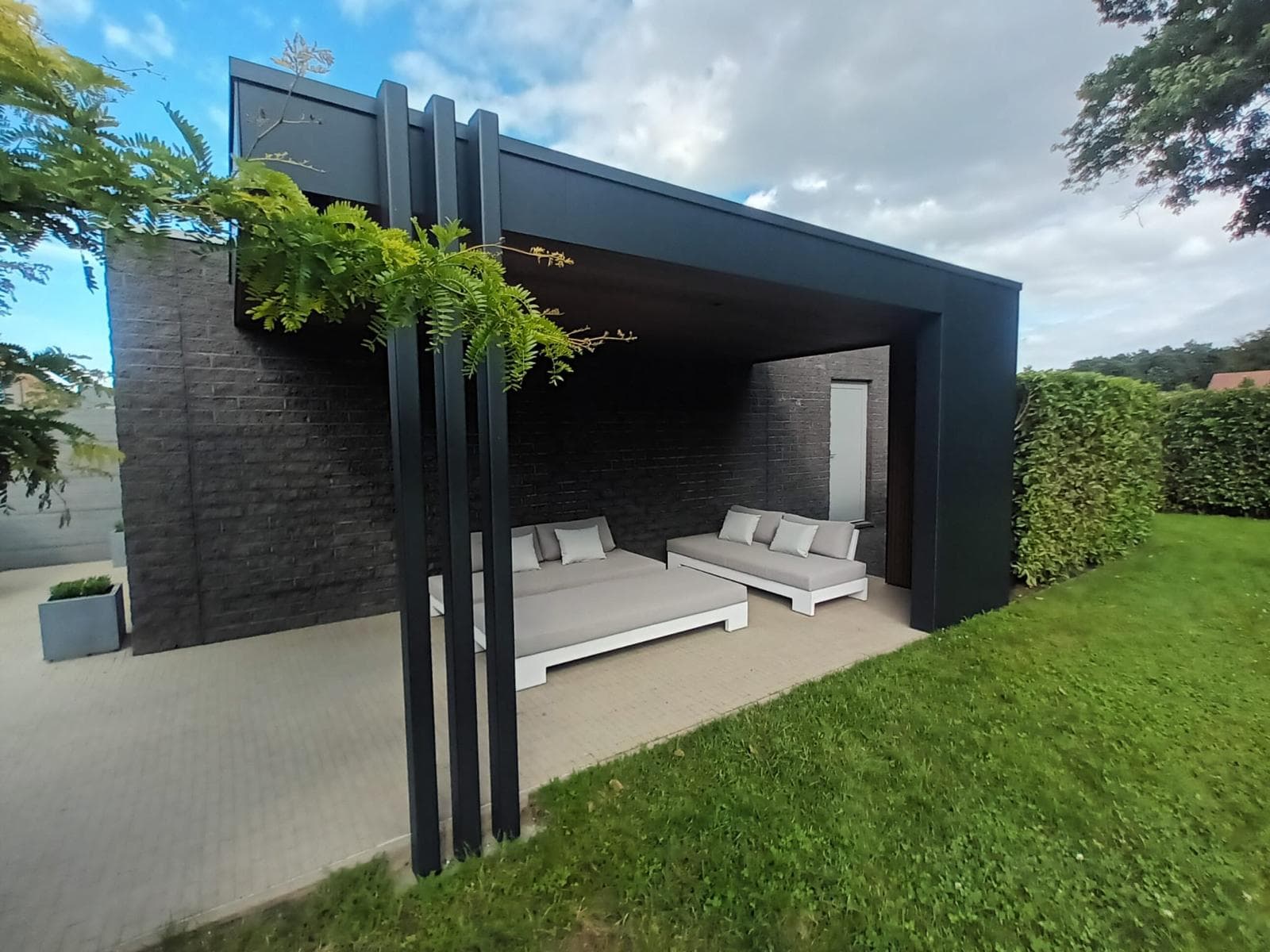 Zomerlounge met twee lichtgrijze daybeds onder de overkapping tegen de zwarte gevelsteen