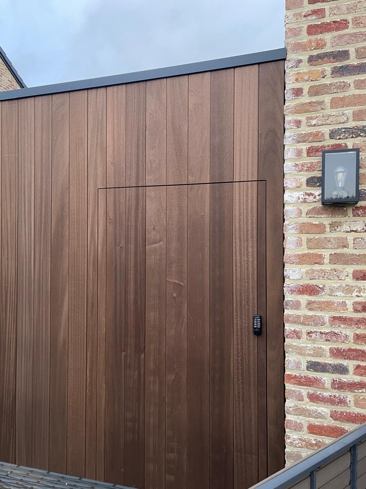 Vlakke houten Ayous-wand met geïntegreerde codetoegangsdeur naast bakstenen gevel