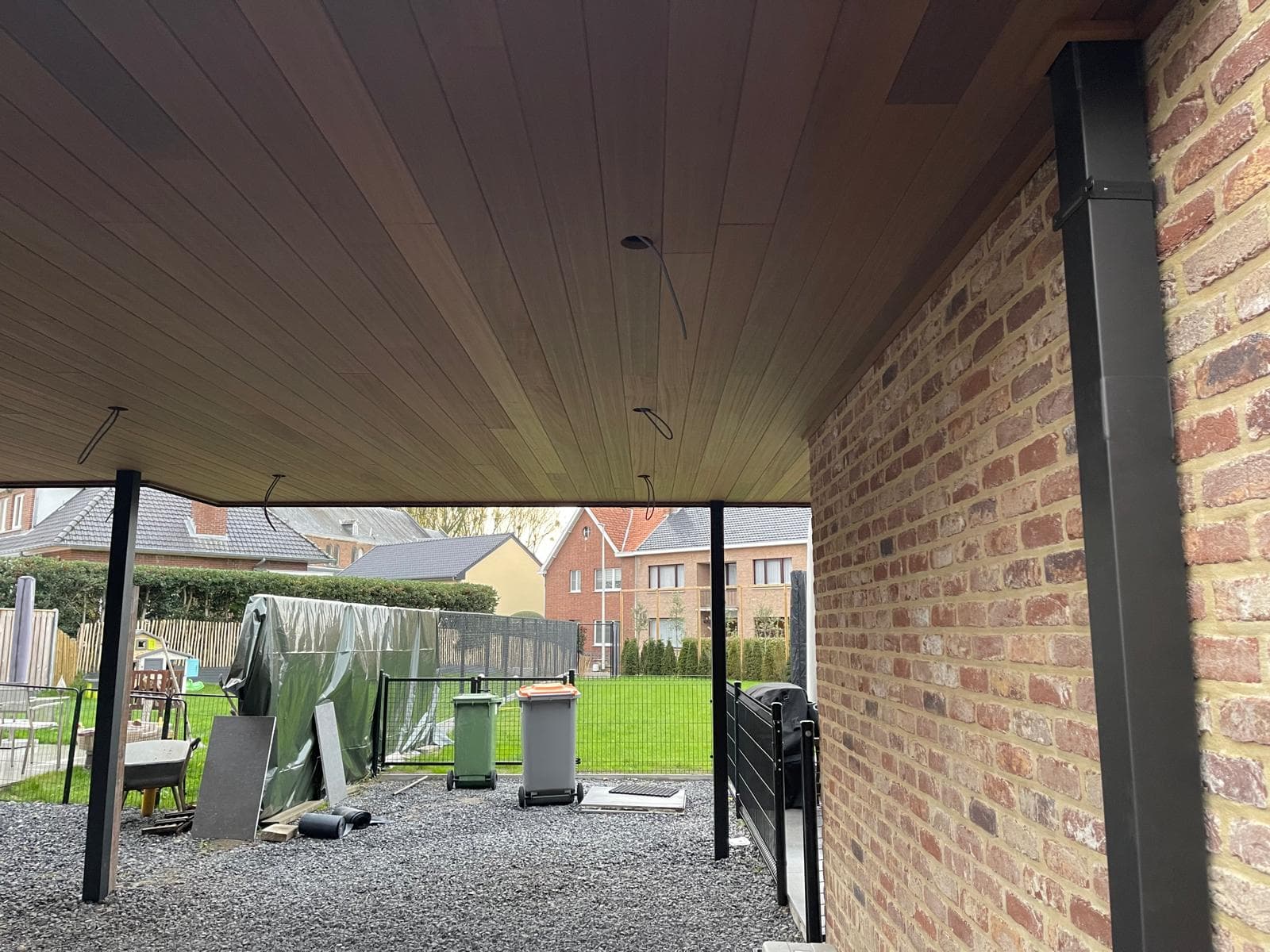 Houten carportplafond dat aansluit op de bakstenen gevel met zwarte stalen kolommen