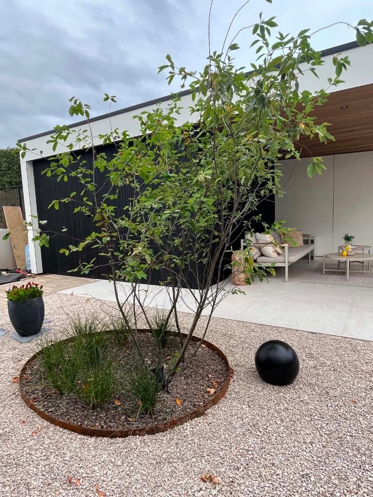 Kiezeltuintje met Corten-rand en zwarte sfeerbol, lounge van de poolhouse op de achtergrond