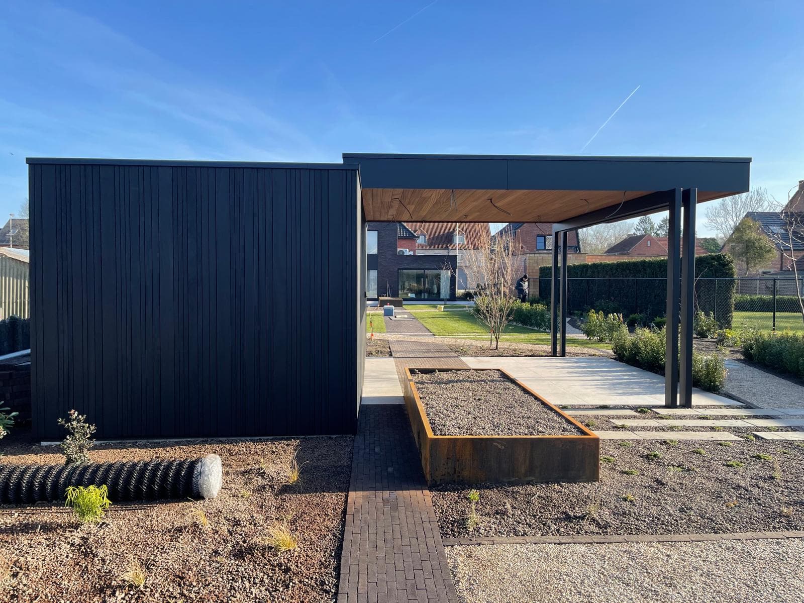 Vooraanzicht van de zwarte tuinberging met overkapping, cortenstalen plantbak en bestraat pad